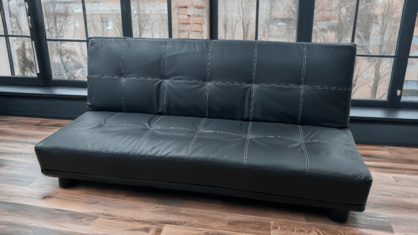 Le clic-clac "Crea", un canapé convertible en simili cuir noir avec des surpiqûres apparentes, allie design moderne et fonctionnalité. Son assise capitonnée offre un confort optimal, tandis que ses pieds discrets assurent une bonne stabilité. Placé dans un intérieur lumineux avec de grandes fenêtres et un parquet en bois, ce canapé apporte une touche contemporaine et élégante, parfait pour un salon ou un espace polyvalent.