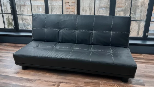 Le clic-clac "Crea", un canapé convertible en simili cuir noir avec des surpiqûres apparentes, allie design moderne et fonctionnalité. Son assise capitonnée offre un confort optimal, tandis que ses pieds discrets assurent une bonne stabilité. Placé dans un intérieur lumineux avec de grandes fenêtres et un parquet en bois, ce canapé apporte une touche contemporaine et élégante, parfait pour un salon ou un espace polyvalent.