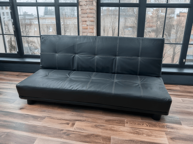 Le clic-clac "Crea", un canapé convertible en simili cuir noir avec des surpiqûres apparentes, allie design moderne et fonctionnalité. Son assise capitonnée offre un confort optimal, tandis que ses pieds discrets assurent une bonne stabilité. Placé dans un intérieur lumineux avec de grandes fenêtres et un parquet en bois, ce canapé apporte une touche contemporaine et élégante, parfait pour un salon ou un espace polyvalent.